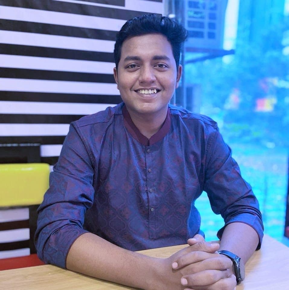নীল নকশা করা পাঞ্জাবি পরা একজন হাসিমুখের যুবক টেবিলে হাত রেখে বসে আছেন।