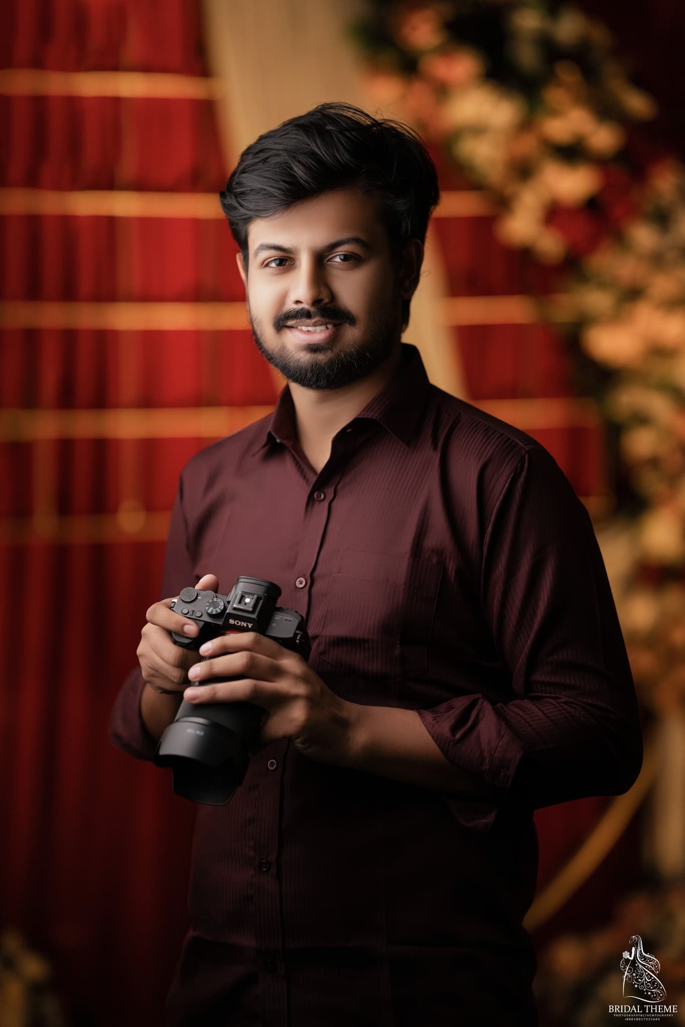 মেরুন শার্ট পরা দাড়িওয়ালা যুবক হাতে একটি ক্যামেরা নিয়ে লাল পর্দার সামনে দাঁড়িয়ে আছেন।